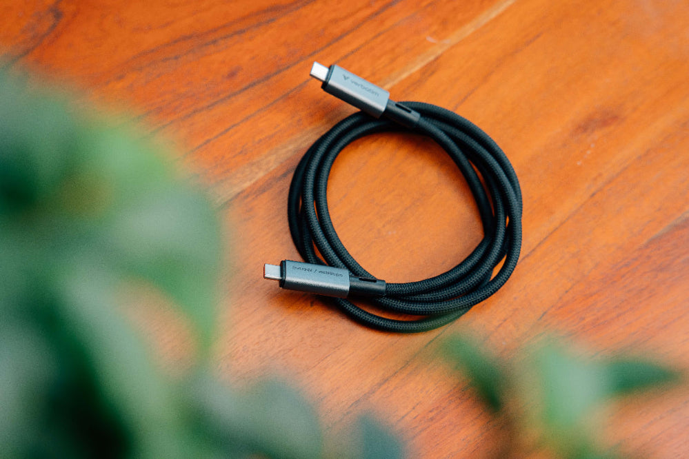 Black USB-C cable on a wooden surface with blurred green plants in the foreground